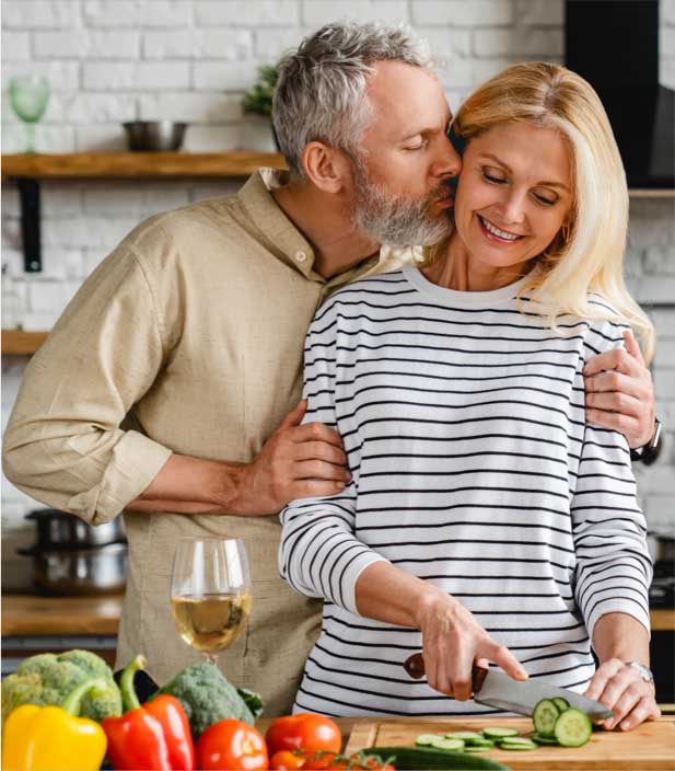 Copeland Tower Living senior apartment in the kitchen couple getting ready to prepping to cook broccoli, bell peppers, tomatoes, and cucumber while husband is hugging smiling wife who is cutting cucumbers and glass of white wine is on the counter
