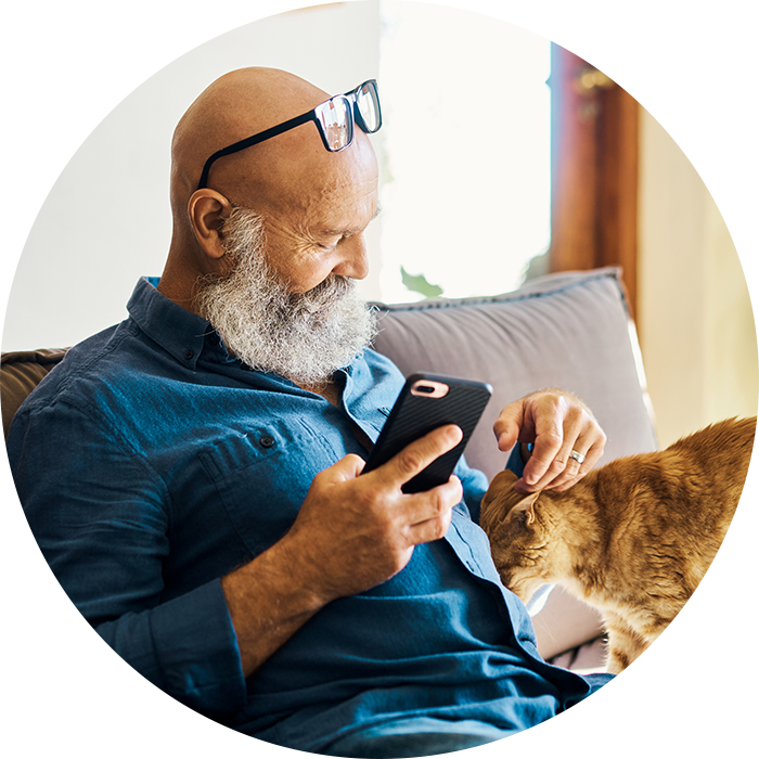 Senior gentleman sitting with glasses on his forehead on the couch smiling while holding a smartphone and petting a orange cat at Copeland Tower Living