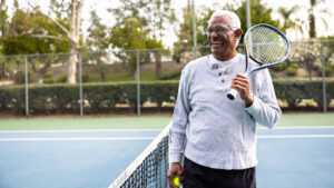 Copeland Tower Living older man on the tennis court holding a tennis ball and tennis racket over his shoulder enjoying one of the best outdoor activities for seniors